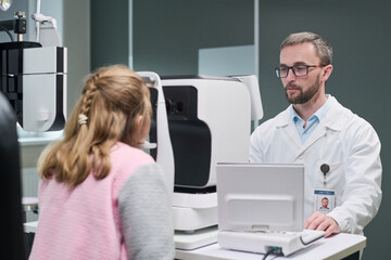 Caucasian middle aged man optometrist conducting eye examination for Caucasian child girl using modern ophthalmic equipment in medical clinic, both focused on diagnostic process