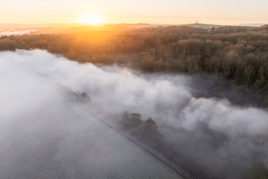 Aerial view of mist rolling over fields and trees, with a distant monument silhouetted against the rising sun, Thames Valley, Oxfordshire, United Kingdom.