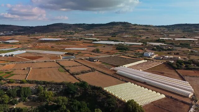 Aerial flyover of agricultural fields, greenhouses, and rural terrain in Mġarr, Malta, capturing the scenic Mediterranean countryside with hills, vegetation, and scattered village structures.