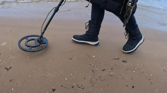 Close-up of a metal detector sweeping across wet coastal sand as boots step forward along the shoreline, capturing treasure-hunting movement and beach.