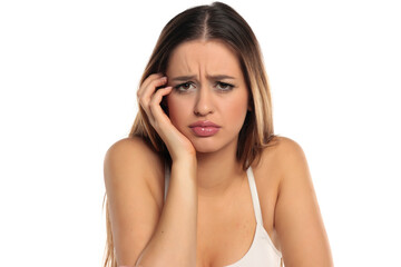 Obraz premium Studio shot of a worried woman with long brown hair and makeup, wearing a white top and resting her head on her hand, on a white background