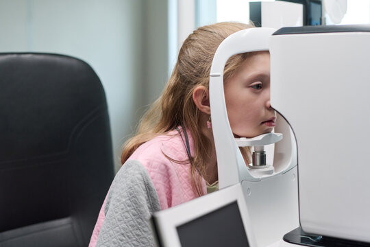 Caucasian girl child undergoing eye examination using ophthalmic diagnostic device in medical clinic, sitting still with face positioned against machine for vision testing - Powered by Adobe