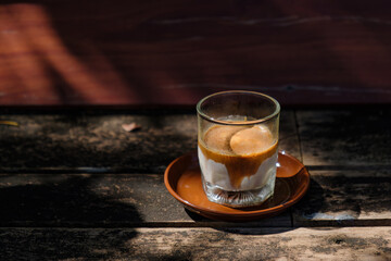 a small shot glass of chilled milk filled with hot espresso shot coffee floated over white creamy chilled milk put on wooden table in wild garden with sun shine and shade of green trees