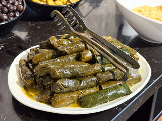 Stuffed dolma with grape leaves arranged on plate with serving tongs. Traditional cuisine, Mediterranean flavors, plant based dish, and cultural food heritage in buffet presentation.