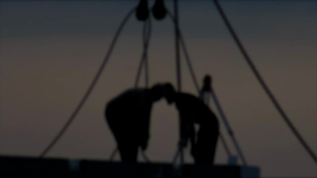 Two workers in silhouette on construction site against twilight sky. Industrial rigging, cables, and pulleys visible. Depicts labor, teamwork, and dedication of people working at dusk.