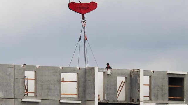 Overcast sky. Construction site. Red crane hook lifts concrete wall panels. Worker guides prefabricated sections. Building structure forms, urban development.