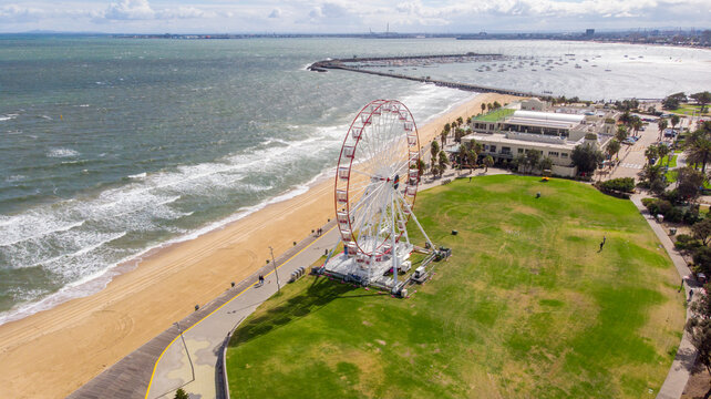 Aerial view of the towering St Kilda Ferris wheel casting shadows on the vibrant green lawn, contrasting with the sandy beach and the vast ocean, St. Kilda, Victoria, Australia.