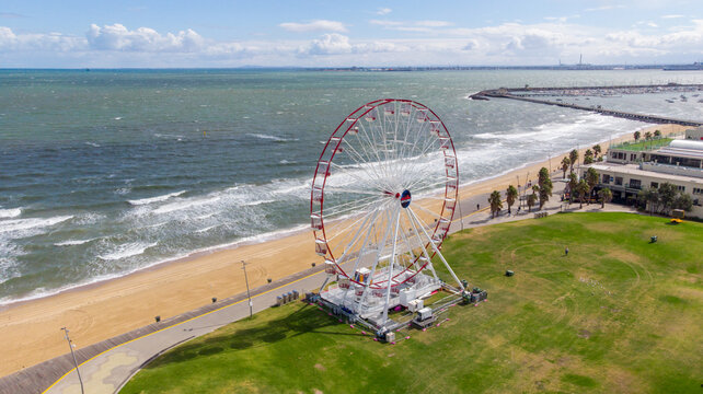 Aerial view of a striking Ferris wheel, its red frame a bold contrast against the soft beach and green grass, under a vast sky, St. Kilda, Victoria, Australia.