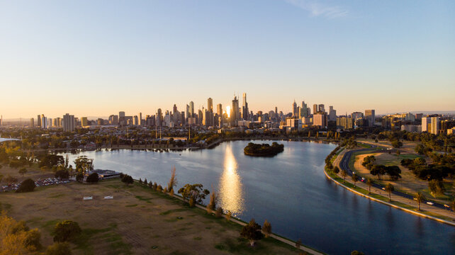Aerial view of the tranquil Albert Park Lake mirroring the golden city skyline under a soft, pastel sky, Albert Park, Victoria, Australia.