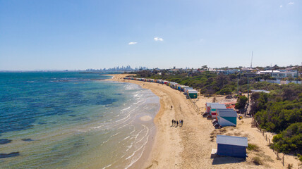 Aerial view of colourful beach boxes line the sandy shore where the turquoise sea meets the land, with the city skyline faintly visible in the distance, Mornington, Victoria, Australia.