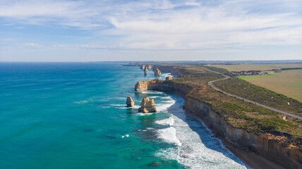Aerial view of rugged cliffs meet the turquoise sea, Twelve Apostles stand tall against the sky, Great Ocean Road winds along the coast, Victoria, Australia.