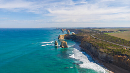 Aerial view of rugged cliffs meet the turquoise ocean, waves crash against the iconic Twelve Apostles, and the Great Ocean Road winds along the coastline, Twelve Apostles, Victoria, Australia.