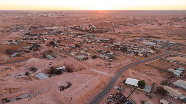 Aerial view of the sun dipping below the horizon, casting long shadows across the arid landscape and small, scattered buildings, Coober Pedy, South Australia, Australia.