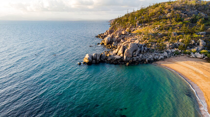 Aerial view of the vivid turquoise waters meeting the golden sands of a secluded beach beneath rocky outcrops and tree-covered headland, Magnetic Island, Queensland, Australia.
