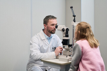 Caucasian middle aged man performing eye examination on Caucasian girl using slit lamp in medical office, doctor focusing on patient while child sitting still during vision test