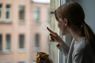 Woman drawing smiley face on foggy window with finger while snow falling outside. Girl makes smile on steamed glass, looking at snowy window holding cup. Winter mood, hope, cozy positive vibe at home