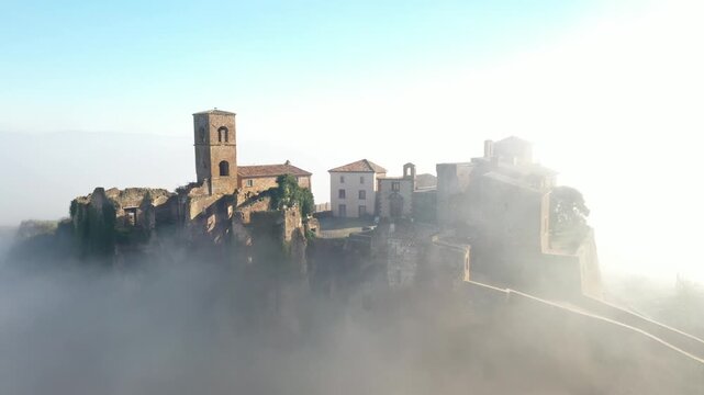 Aerial view of Celleno's historic buildings and tower rising above the mist, with warm stone textures contrasting with the soft, ethereal fog, Celleno, Lazio, Italy.