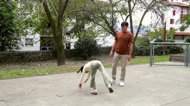 Happy young girl practicing acrobatics, successfully performing a perfect cartwheel on a concrete court while her supportive father proudly watches her during a fun day together outdoors in a park