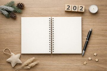 Overhead view of an open blank notebook with a pen, surrounded by winter and New Year's themed decorations like pine cones, a candle, and '2022' blocks on a wooden table.