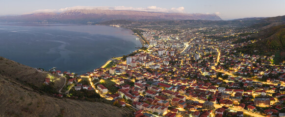 Twilight aerial view of Pogradec, Albania, with illuminated streets, dense city neighbourhoods, and the calm waters of Lake Ohrid at dusk.