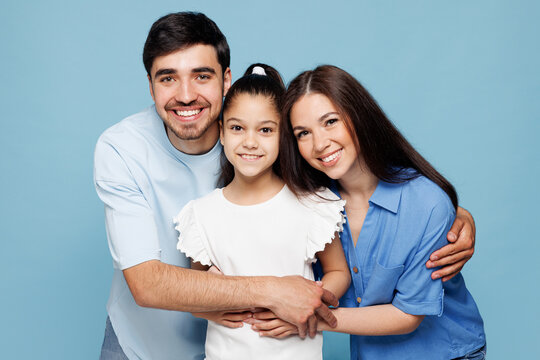 Young smiling cheerful happy parents mom dad with child kid daughter girl wear casual clothes hug embrace posing look camera isolated on plain blue background Family day parenthood childhood concept.