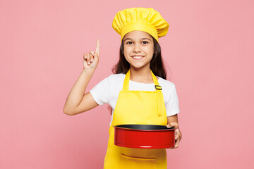 Small kid girl housewife chef cook baker mother's helper wear yellow apron toque cap hold metal baking form for pie, baking dish point finger up isolated on plain pink background Cooking food concept