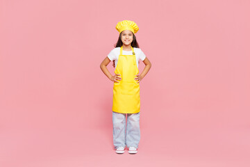 Full body smiling happy small kid child girl housewife chef cook baker mother's helper wearing yellow apron toque cap look camera stand akimbo isolated on plain pink background. Cooking food concept.