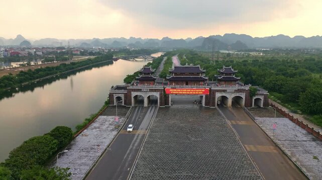 Aerial view of the traditional gate casting a shadow on the road with a car passing through, alongside the river reflecting the sky, Hoa Lu, Ninh Binh, Vietnam.