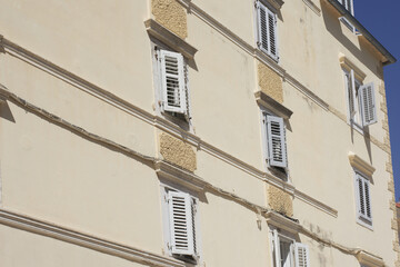 Two simple, square windows with white frames set into a weathered, rustic stone and plaster wall of an old European building under a terracotta roof. © Impresyjna
