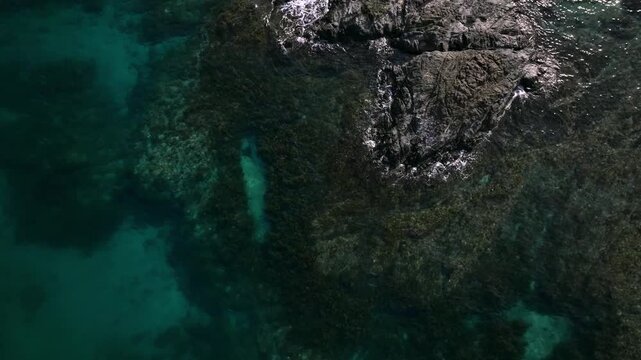 Aerial view of a dark rocky outcrop surrounded by turquoise sea, creating a stark contrast in tones and textures, Carles, Western Visayas, Philippines.