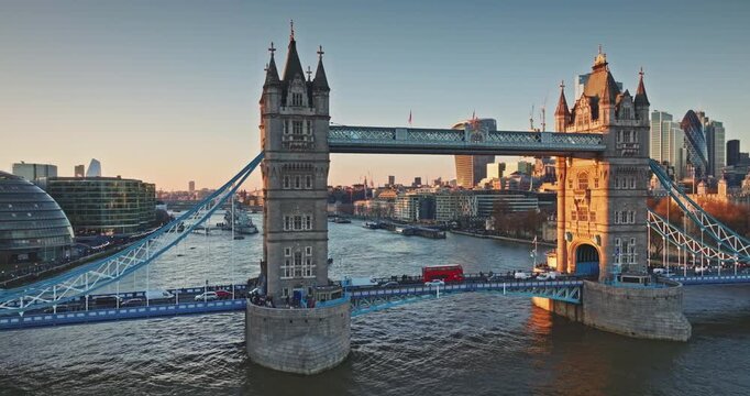 Tower Bridge Victorian Gothic towers and bustling cars traffic cross the River Thames at golden hour sunset, London skyline glowing in warm evening light. Travel destination. Aerial panorama footage
