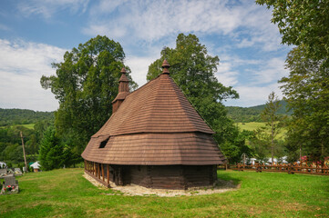 View of aged wooden church with its dark brown shingled roof standing against the backdrop of lush green trees and a blue sky, Topola, Presovsky kraj, Slovakia.