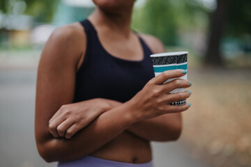 A woman in a black sporty top stands outdoors with arms crossed, holding a patterned disposable cup. The scene conveys a casual, active lifestyle and hydration during outdoor activity.