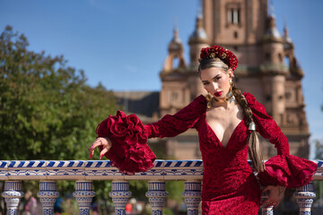 Obraz premium Young, beautiful, blonde woman in red flamenco dress, flowers in her hair and gold jewelry, performing flamenco poses leaning on a tiled railing in the square of Spain in Seville. Dance concept, art.