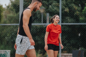 A man in a black tank top and patterned shorts and a woman in a bright red shirt and black shorts share a light moment during an outdoor training session on a fenced court.