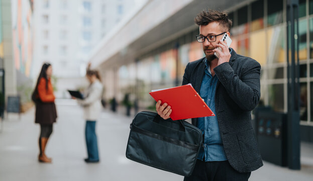 A stylish man in a blazer uses a mobile phone while carrying a red folder and a black briefcase in a modern urban setting, with blurred pedestrians in the background.