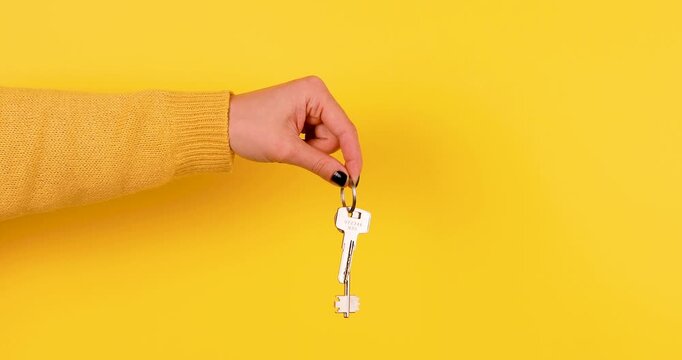 Close-up of a female hand holding house keys against a bright yellow background. Minimal 4K video ideal for real estate, home ownership, rental, moving, security, and property concepts.