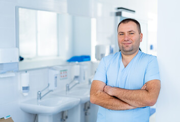 Obraz premium Confident Medical Specialist Standing with Crossed Arms. Waist-up portrait of a friendly male doctor in blue scrubs standing with arms crossed in a hospital examination room with a sink.