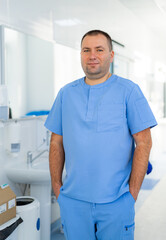 Fototapeta premium Portrait of Confident Male Doctor in Blue Uniform in Hospital. Portrait of a professional male doctor wearing blue medical scrubs standing with hands in pockets in a modern clinic hallway.