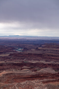 View of layered red rock canyons carved by winding green rivers under a vast, cloudy sky, creating a dramatic, textured landscape, Moab, Utah, United States.