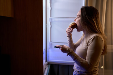 Female eating muffin beside open refrigerator in kitchen at night