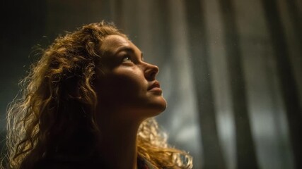 A woman with curly hair gazes upward, illuminated from above against a dark, moody background.
