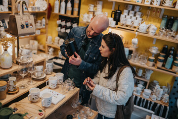 A couple browses a colorful pottery shop, studying mugs and tea wares together. They discuss items and pick a few as gifts in a cozy store setting.