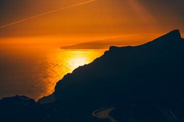 Stunning sunset over the ocean from the Masca village area in Tenerife, with the silhouette of La Gomera island visible in distance. Sky is a brilliant orange hue, reflecting brightly on water