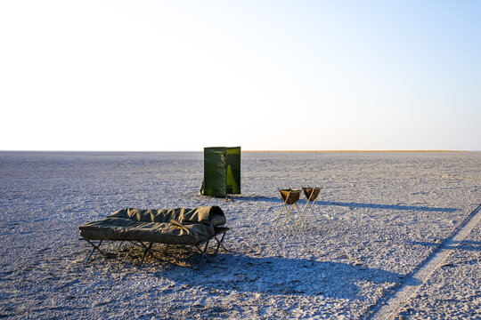 Minimalist camp setup on the Makgadikgadi Pan, Botswana. Bed, chairs, and outdoor shower on endless salt flats, capturing remote luxury and solitude in Africa.