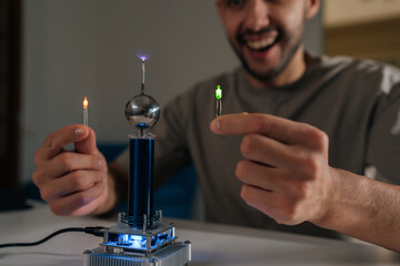 Excited young man demonstrating tesla coil wireless power transmission, illuminating two light bulbs without direct contact, showcasing electricity, science and hobby experiment, close-up.