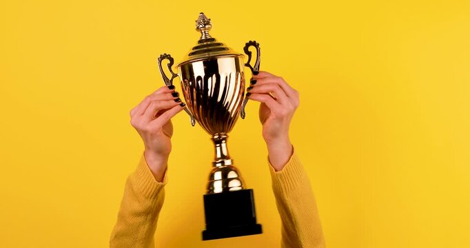 Close-up of a female hands holding a golden sports trophy cup on a bright yellow background. Clean, minimal 4K video ideal for victory, success, achievement, competition, and award concepts.