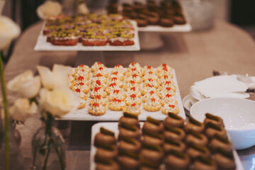Several platters hold small desserts at a gathering. Treats include cream-filled pastries and cookies. White flowers add a delicate touch to the scene. Guests enjoy the sweet offerings and socialize