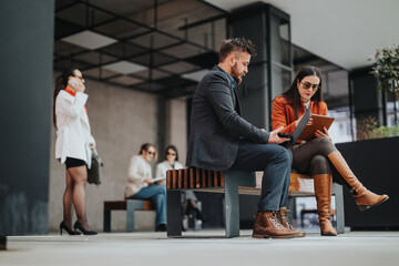 A man and a woman sit on a wooden bench, examining a laptop and notebook, while colleagues chat and use their phones nearby in a contemporary, open-air workspace.