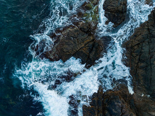 Waves crash dramatically against rugged rocks at the coastline under a bright blue sky, creating a...